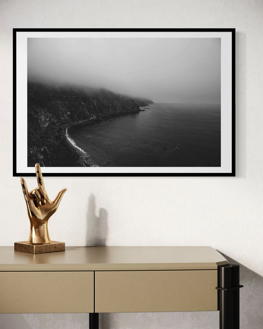 Framed black and white landscape photo print of a coastal scene on a wall above a desk with a gold hand sculpture.